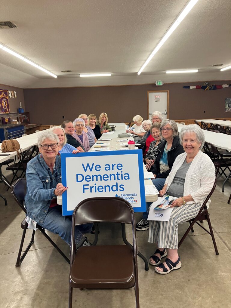 Group Photo of Senior citizens siting at a table. One old lady on the front left is holding a blue sign saying, 'We are Dementia Friends'